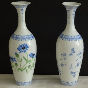 Può includere: Due vasi in porcellana bianca con motivi floreali blu e verdi. Il vaso a sinistra ha un motivo floreale più grande con fiordalisi blu e foglie verdi. Il vaso a destra ha un motivo floreale più piccolo con fiori blu e foglie verdi.