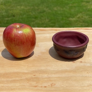 May include: A red and yellow apple next to a small, maroon-colored ceramic bowl. The apple has a shiny surface and is sitting on a light brown wooden surface. The bowl has a textured exterior.