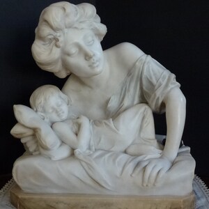 May include: A white marble sculpture of a woman cradling a sleeping child. The woman has long hair and is wearing a simple robe. The child is lying on a pillow and is covered with a blanket.