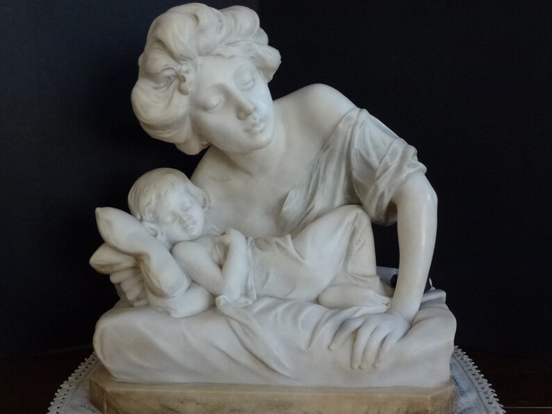 May include: A white marble sculpture of a woman cradling a sleeping child. The woman has long hair and is wearing a simple robe. The child is lying on a pillow and is covered with a blanket.