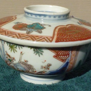 Könnte beinhalten: Eine weiße Porzellanschüssel mit Deckel, verziert mit einem floralen und geometrischen Muster in Rot, Blau und Gold. Die Schüssel ist rund und hat einen kleinen Fuß.