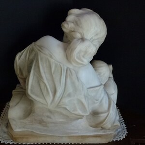 May include: A white marble sculpture of a woman holding a child. The woman is seated and the child is resting on her lap. The sculpture is on a white base.