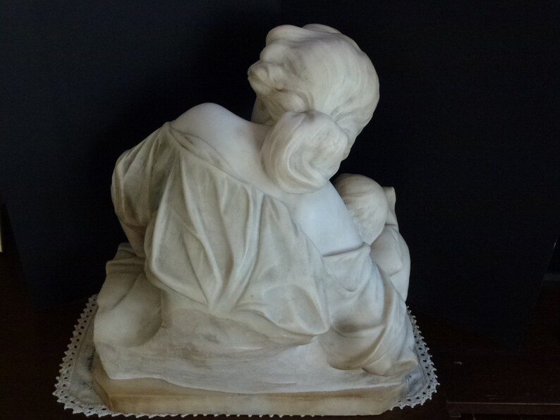 May include: A white marble sculpture of a woman holding a child. The woman is seated and the child is resting on her lap. The sculpture is on a white base.
