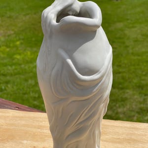 Op de afbeelding: Een witte keramische vaas met een gebeeldhouwd ontwerp van twee figuren die elkaar omarmen. De vaas heeft een gladde, matte afwerking en een klassieke, elegante vorm. De figuren zijn gehuld in vloeiende stof, wat een gevoel van beweging en gratie aan het stuk toevoegt.