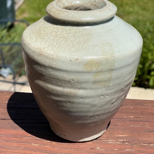 Könnte beinhalten: Eine hellgrüne Keramikvase mit breitem Körper und schmalem Hals. Die Vase hat eine strukturierte Oberfläche mit sichtbaren Rissen und horizontalen Rillen. Die Öffnung ist in einem dunkleren Grünton gehalten.