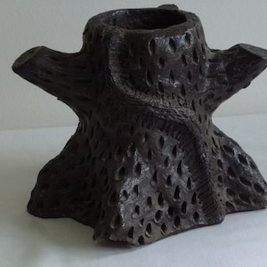 Puede incluir: Un jarrón de cerámica marrón oscuro con forma de tronco de árbol con una superficie rugosa y texturizada y un centro hueco.