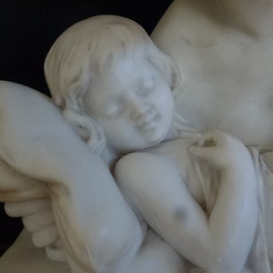 May include: A white marble sculpture of a mother holding a sleeping child. The child is resting its head on a pillow and has its arm around the mother's neck.