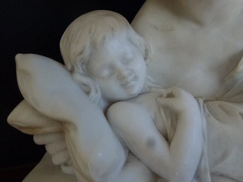 May include: A white marble sculpture of a mother holding a sleeping child. The child is resting its head on a pillow and has its arm around the mother's neck.
