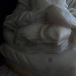 May include: White marble sculpture of a person sitting with their arms crossed and holding a pillow.