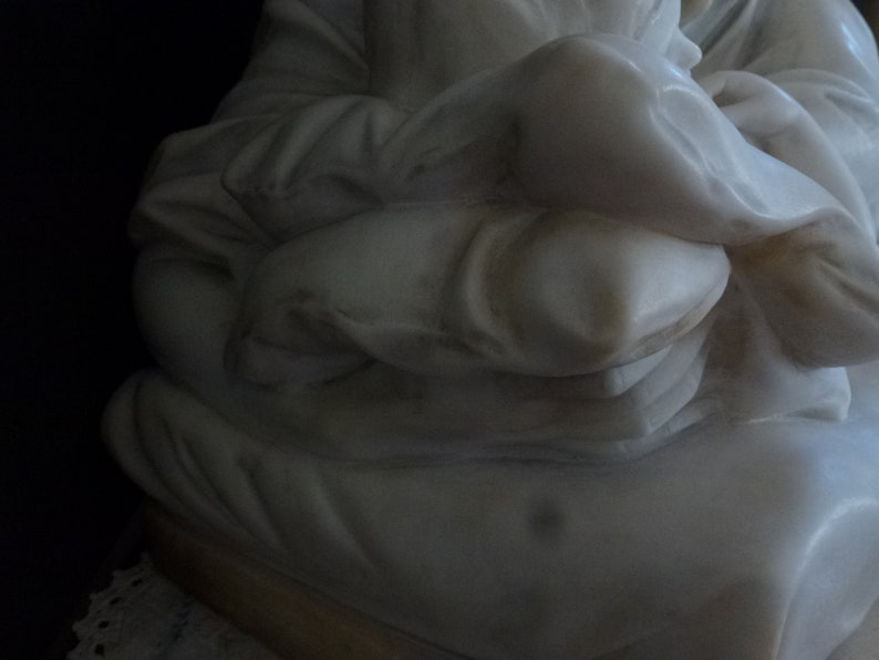 May include: White marble sculpture of a person sitting with their arms crossed and holding a pillow.