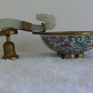 May include: A small, white porcelain bowl with a floral design in shades of green, blue, purple, and orange. The bowl is on a brass stand with a jade handle in the shape of a dragon's head.