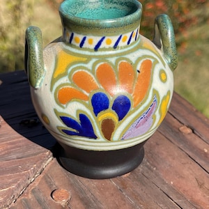 Może przedstawiać: Mały, ceramiczny wazon z okrągłym korpusem i dwoma uchwytami. Wazon ma kremową podstawę z kwiatowym wzorem w kolorze pomarańczowym, niebieskim i żółtym. Obręcz i uchwyty są zielone, a podstawa czarna. Wazon ma około 10 cm wysokości.