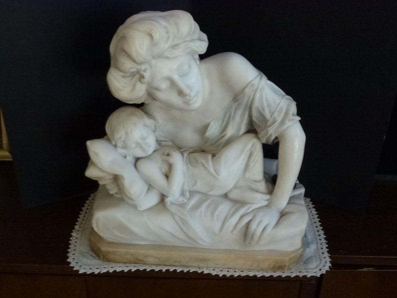 May include: A white marble sculpture of a woman holding a sleeping child. The woman is sitting and the child is lying on her lap. The sculpture is on a white base with a lace trim.