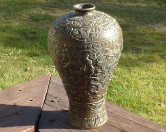May include: Bronze-colored decorative vase with intricate relief carvings. The vase features detailed depictions of dragons and floral patterns. It stands on a wooden surface against a backdrop of green grass.