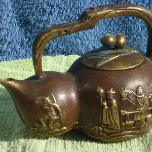 May include: A small, brown, bronze teapot with a carved handle and lid. The teapot is decorated with raised figures of people and animals.