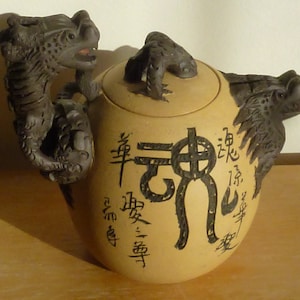 May include: A tan ceramic teapot with a dragon-shaped handle and spout. The lid features a small dragon figure. Black calligraphy decorates the body of the teapot. The teapot is a unique piece of Asian-inspired decor.