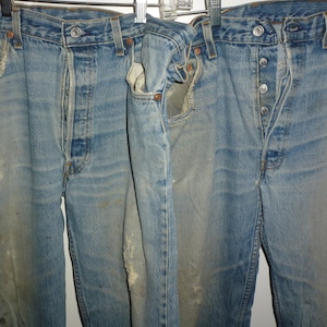 Puede incluir: Tres pares de pantalones vaqueros azules de mezclilla con varios detalles desgastados, que incluyen roturas y áreas descoloridas. Los vaqueros están colgados en un tendedero.