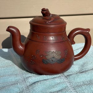 May include: A reddish-brown ceramic teapot with a frog-shaped lid handle. The teapot features a curved spout and handle, with a lotus flower design on the body. The teapot is sitting on a blue towel.