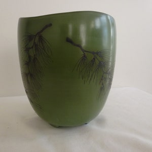 May include: A green ceramic vase with a black pine branch design. The vase has a rounded shape and a wide opening.