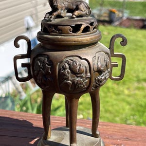Puede incluir: Quemador de incienso de bronce con un diseño detallado. El quemador presenta una tapa con una pequeña figura de animal, asas ornamentadas y tallas en relieve de pájaros y otras figuras. El quemador se apoya en tres patas.