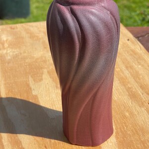 May include: A tall, burgundy-colored vase with a textured, swirling design. The vase has a wide opening and a hexagonal base. The color gradient transitions from a darker shade at the top to a lighter shade at the bottom. The vase is made of ceramic.