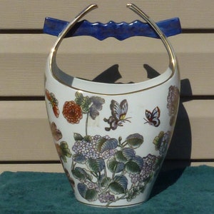 Op de afbeelding: Een witte porseleinen vaas met een blauw en gouden handvat. De vaas is versierd met een bloemenpatroon met vlinders, bloemen en bladeren.