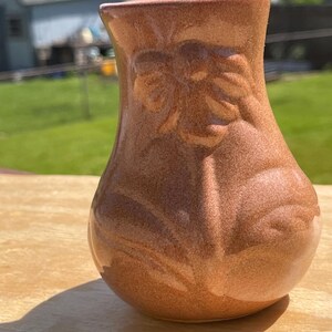 May include: A small, brown ceramic vase with a textured surface and embossed floral design. The vase has a rounded body and a slightly flared top. The vase is sitting on a wooden surface.