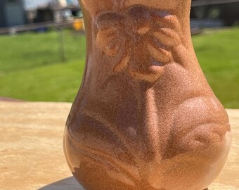 May include: A small, brown ceramic vase with a textured surface and embossed floral design. The vase has a rounded body and a slightly flared top. The vase is sitting on a wooden surface.