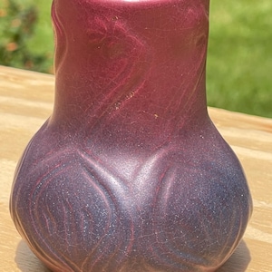 Könnte beinhalten: Eine handgefertigte, dekorative Vase mit rundem Boden und schmalem Hals. Die Vase hat einen Farbverlauf, der von einem tiefen Lila am Boden zu einem rötlichen Lila am oberen Ende übergeht. Die Oberfläche hat ein strukturiertes, craquelé Aussehen.