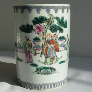Pode incluir: Um vaso cilíndrico de porcelana branca com uma borda azul e dourada. O vaso é decorado com uma cena pintada à mão de três figuras em uma paisagem com um pinheiro e uma flor rosa.