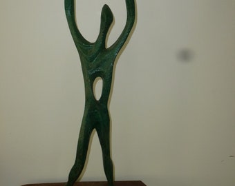 Può includere: Scultura astratta di una persona con le braccia alzate, realizzata in metallo verde e montata su una base in legno.