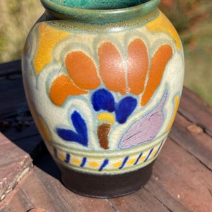 Może przedstawiać: Mały, okrągły wazon ceramiczny z zieloną obwódką i ciemnobrązową podstawą. Wazon ma kwiatowy wzór w kolorze pomarańczowym, niebieskim i żółtym na kremowym tle. Wazon stoi na drewnianej powierzchni.