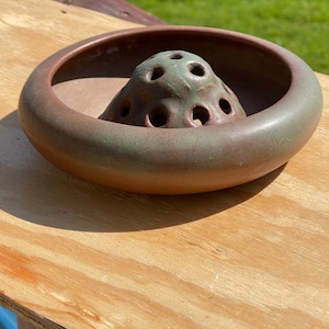 May include: A ceramic flower frog with a wide, round rim in shades of brown and green. The center features a cone-shaped structure with multiple holes for arranging flowers. The frog is designed to hold stems in place.