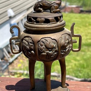 May include: A bronze incense burner with a detailed design. The burner features a lid with a small animal figure on top, three legs, and handles on the sides. The body of the burner has relief carvings of figures and floral patterns.