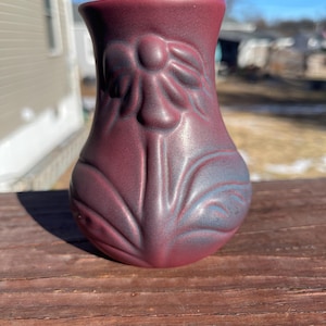 May include: A maroon-colored ceramic vase with a floral design. The vase has a bulbous base and a flared rim. The floral design is embossed on the surface of the vase. The vase is sitting on a wooden surface.
