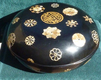 May include: A black lacquered box with a round lid decorated with gold and mother-of-pearl inlays. The lid features a variety of floral and geometric designs, including a central Chinese character.
