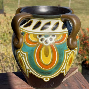 May include: A decorative ceramic vase with a dark brown interior and handles. The exterior features a teal, orange, yellow, and white floral design. The vase is sitting on a wooden surface.