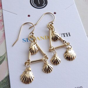 May include: Gold triangle earrings with four dangling seashell charms. The earrings are on a white card with the text "Sea and South" and a website address.