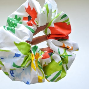 Pode incluir: Uma grande scrunchie floral em tecido branco com um padrão colorido de flores e folhas laranja, verdes, amarelas e azuis. A scrunchie é segurada em uma mão, mostrando seu design volumoso e franzido.