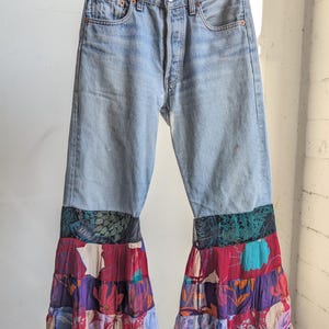 Puede incluir: Vaqueros azul claro con bajos acampanados de patchwork. El patchwork presenta estampados florales y abstractos en tonos rojos, morados y negros. Los vaqueros cuelgan de una percha de madera.