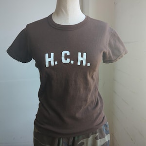 Puede incluir: Una camiseta marrón con las letras blancas "H.C.H." impresas en el pecho. La camiseta se muestra en un maniquí y se combina con pantalones cortos de camuflaje. La camiseta tiene mangas cortas y cuello redondo.