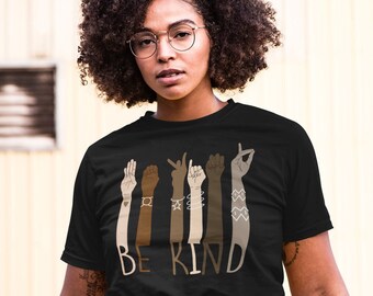 May include: Black t-shirt with the words "BE KIND" and illustrations of hands in various skin tones. The hands are making the sign language symbol for "kind". The shirt is worn by a person wearing glasses and blue jeans.