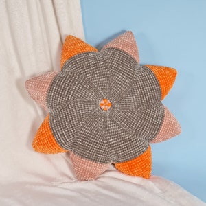 May include: A plush star-shaped cushion with a grey centre and orange and pink accents. The cushion has a textured surface and a small flower-shaped button in the centre.
