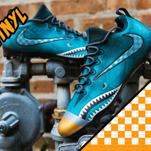 Puede incluir: Botas de fútbol azul turquesa y negras con la punta dorada y un diseño de dientes de tiburón. El swoosh de Nike es blanco con un patrón de cuadros negros y blancos.