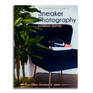 May include: A book titled "A Customizer's Guide to Sneaker Photography, Extended Edition" is displayed. A navy blue and coral sneaker with a white sole rests on a dark blue velvet chair. The book's authors are Dillon DeJesus & Jason.