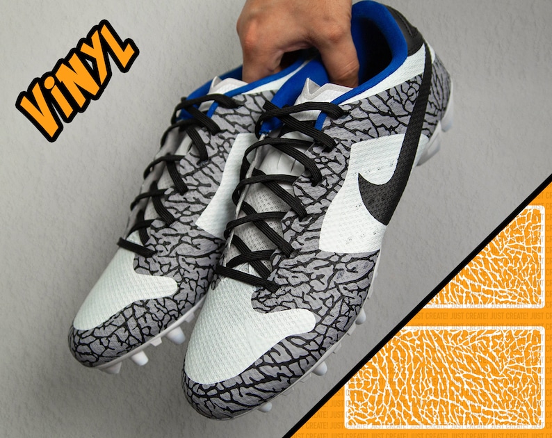 May include: A pair of white soccer cleats with a black and gray elephant print design. The cleats have black laces and a black Nike swoosh logo. The word "VINYL" is written in orange letters on the background.