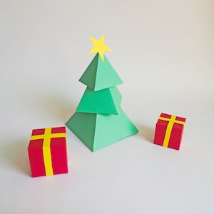Op de afbeelding: Een groene papieren kerstboom met een gele ster bovenop en twee rode cadeaudozen met een geel lint.