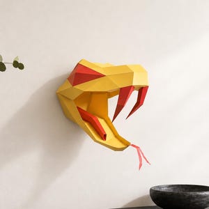 Puede incluir: Una escultura de pared con forma de cabeza de serpiente geométrica amarilla y roja. La escultura está montada en una pared blanca. Un jarrón con vegetación y un cuenco oscuro están sobre una superficie gris oscuro debajo.