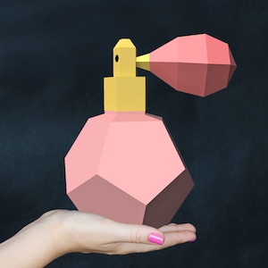 Puede incluir: Un frasco de perfume geométrico rosa con una tapa amarilla y un pulverizador rosa. El frasco está siendo sostenido en una mano.