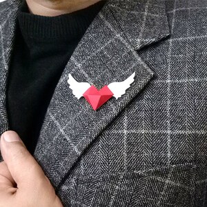 Könnte beinhalten: Ein rotes Herz mit weißen Flügeln, ein geometrisches Papierdesign, ist an einem grauen karierten Blazer befestigt.